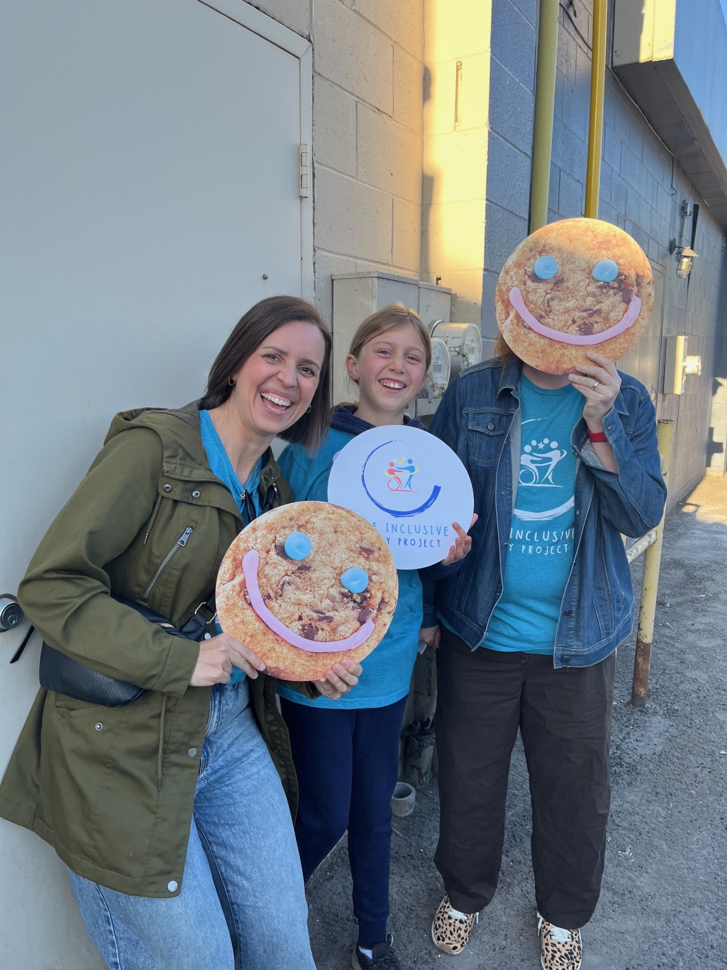 Photo of Inclusive play project volunteers holding smile cookie signs and a sign with the inclusive play project logo while smiling.