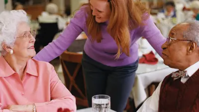 Two older people at dinner being greeted by a worker