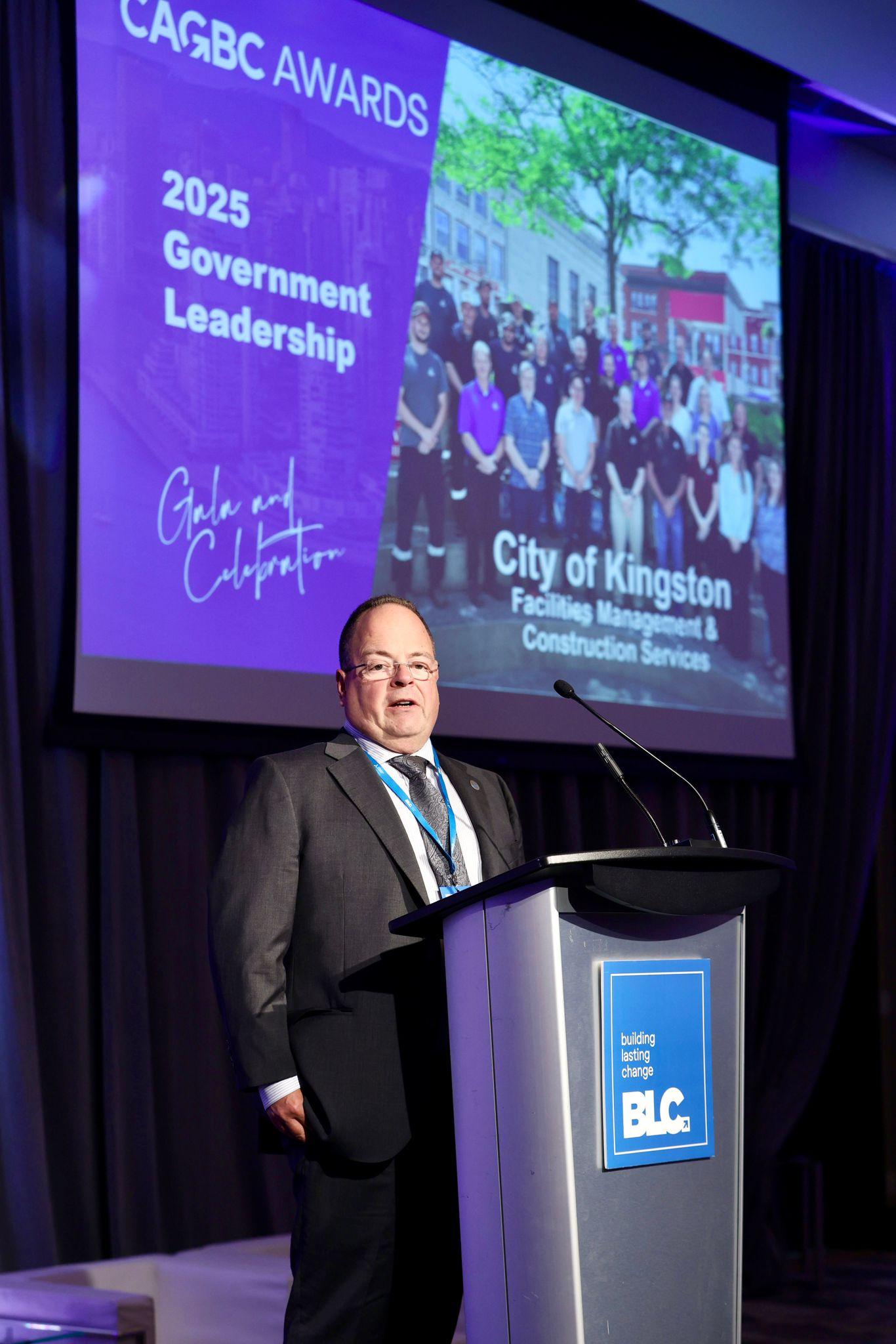 Speros Kanellos stands at a podium with a screen reading 2025 Government Leadership and City of Kingston Facilities Management & Construction Services in the background.
