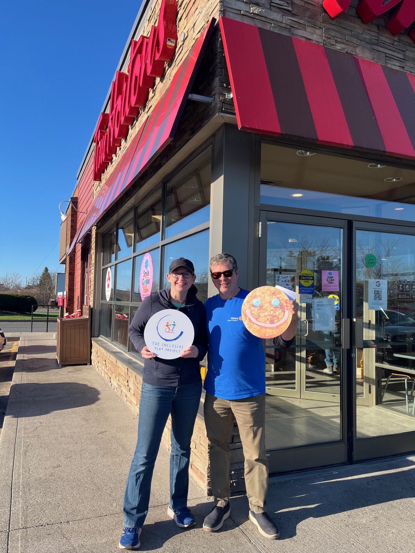 Two people standing outside Tim Hortons holing one circular sign with the inclusive play project logo and a sign with a smile cookie on it.