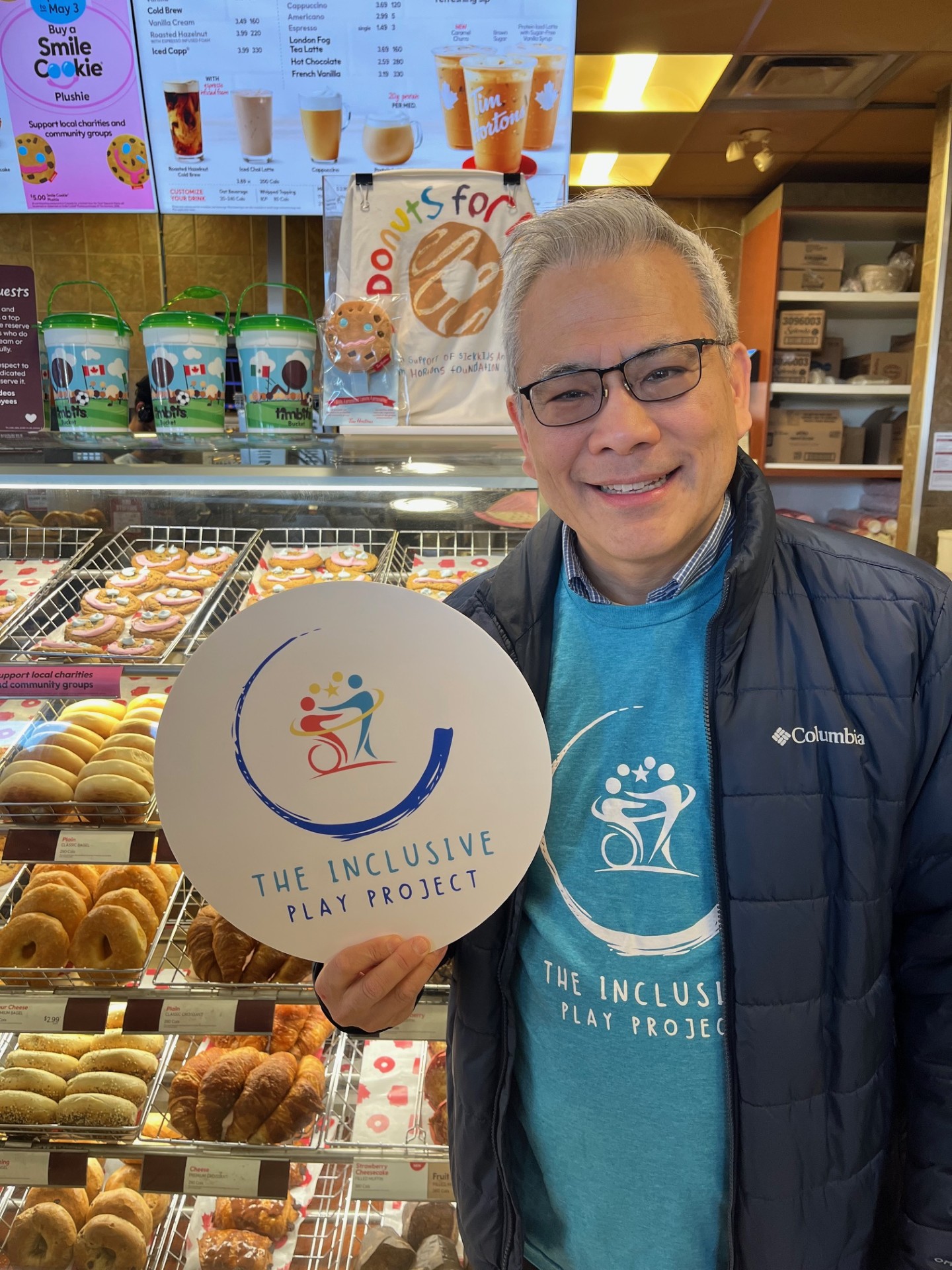 Man standing in front of Tim Hortons donut display holding a circular sign with the inclusive play project logo on it.