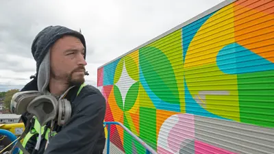 Painter painting the large mural on the side of a building while utilizing a cherry picker